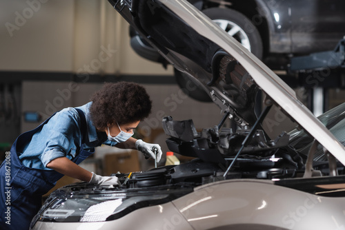 young african american mechanic in protective mask holding wrench and working with car motor in garage