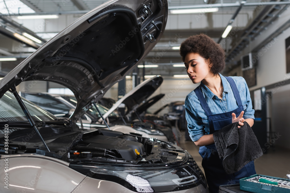 young african american mechanic drying hands with towel near car with open hood in garage young african american mechanic drying hands with towel near car with open hood in garage