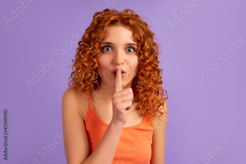 Cute red-haired girl with curls holds her finger to her lips, speaking softly, shh isolated on purple background.