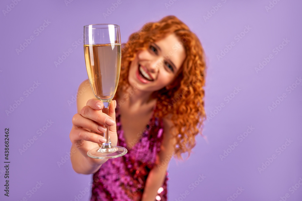 Cute red-haired woman with curls in a shiny dress and a glass of champagne is having fun isolated on a purple background. The concept of a party, celebration, birthday. Cute red-haired woman with curls in a shiny dress and a glass of champagne is having fun isolated on a purple background. The concept of a party, celebration, birthday.