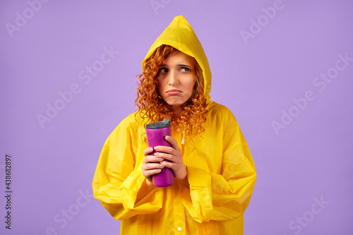 Redhead woman in yellow raincoat and thermal mug isolated on purple background.