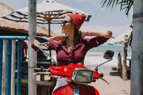 Close up portrait of stylish lady dressed cap and shirt and sunglasses sitting on red bike and smiling on background of ocean