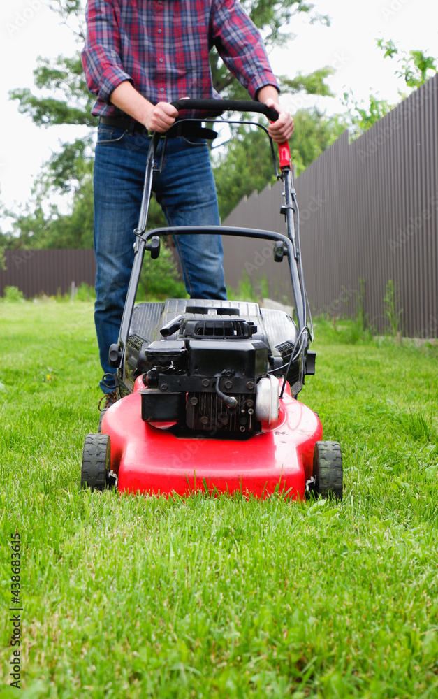 Lawn grass mowing. A man in a plaid shirt and blue jeans mows the grass with a lawn mower. Lawn grass mowing. A man in a plaid shirt and blue jeans mows the grass with a lawn mower.