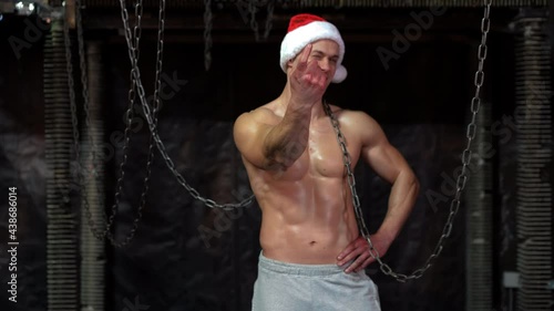 Christmas. Athletic caucasian guy Santa hat pointing finger in cameraYoung shirtless muscular man standing among metal chains, looking at camera photo