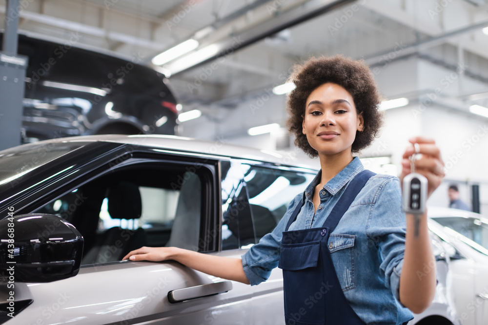 smiling young african american mechanic in overalls holding key near car in garage smiling young african american mechanic in overalls holding key near car in garage