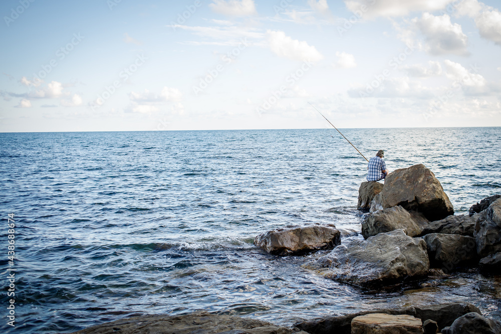 A fisherman is fishing in the sea sitting on large boulders.