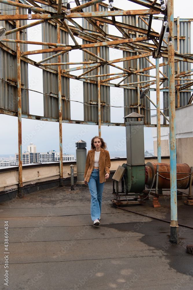 full length of blonde woman in denim jeans and stylish sneakers walking on rooftop full length of blonde woman in denim jeans and stylish sneakers walking on rooftop