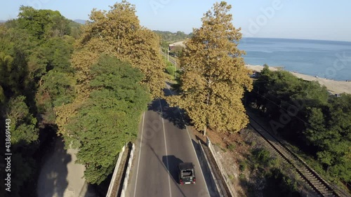 Black suv automobile drives along grey asphalt coastal road between green forest and endless blue sea on sunny summer day aerial follow view photo