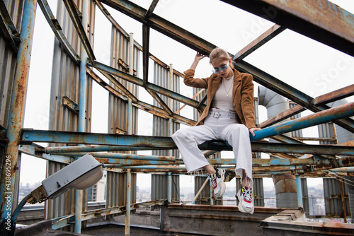 young woman in blue sunglasses and trendy outfit sitting on rusty construction of rooftop