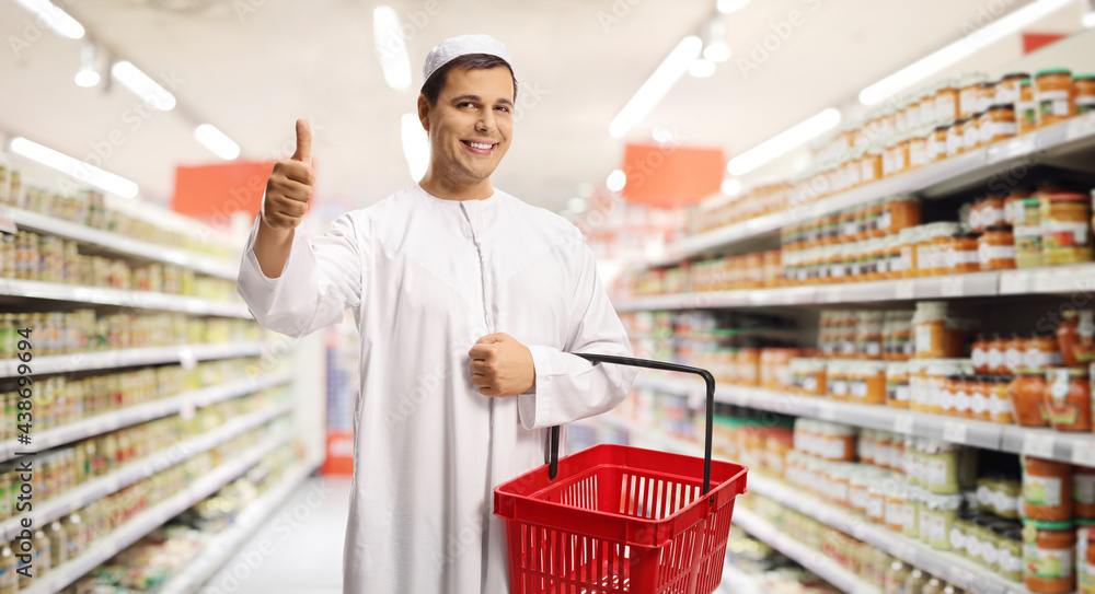 Young man in ethnic clothes holding an empty shopping basket and gesturing a thumb up sign in a supermarket