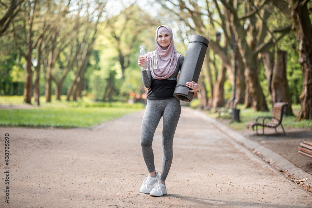 Full length portrait of beautiful muslim woman with yoga mat in hand drinking water after training on fresh air. Pleasant young female wearing sport clothes and hijab.