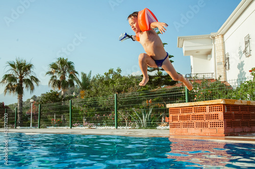 Cute little child boy playing in outdoor swimming pool on hot summer day. Kid jumping in pool. Happy child with orange floaties. Swimming aid protection for kid. Family summer vacation.
