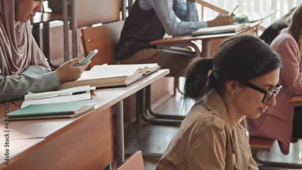 Tilt-up medium slowmo of young multi-ethnic university or high school students sitting in rows by long wooden desks in auditorium and scrolling through smartphones Tilt-up medium slowmo of young multi-ethnic university or high school students sitting in rows by long wooden desks in auditorium and scrolling through smartphones