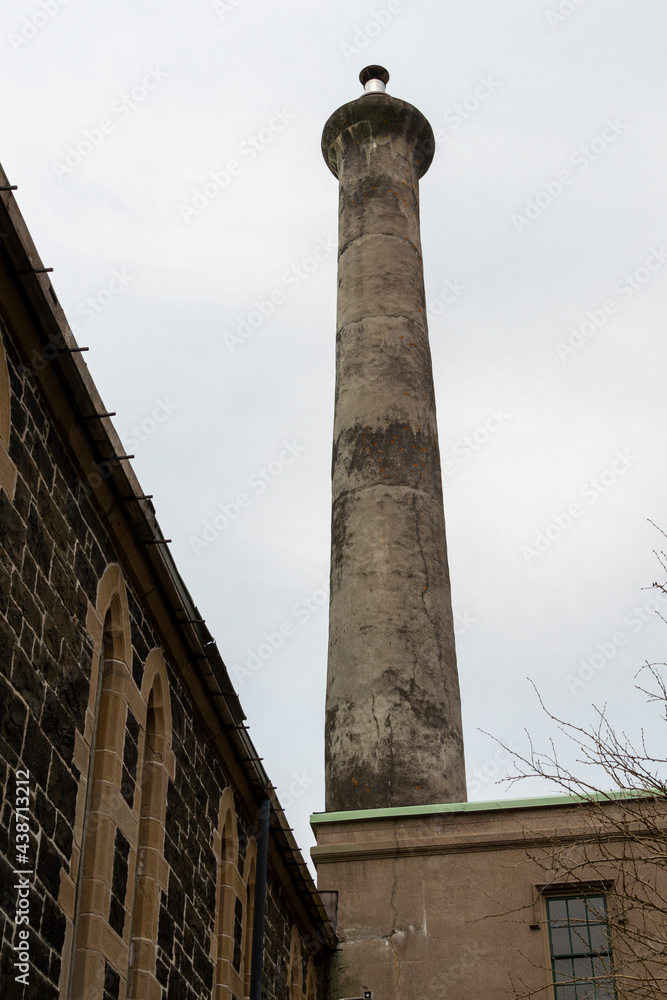 A tall circular smoke stack made of concrete with a  wide cap. The oil furnace smokestack is between two brick buildings. One is a church and the other a vintage office building. The sky is cloudy.