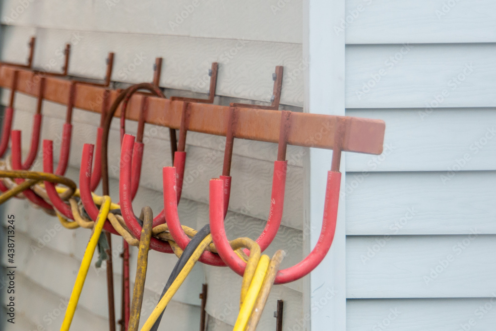 Multiple red plastic hooks on a long metal holder. The hooks have yellow and black extension cords and electrical cords. The holder is attached to a white building that is covered in siding.