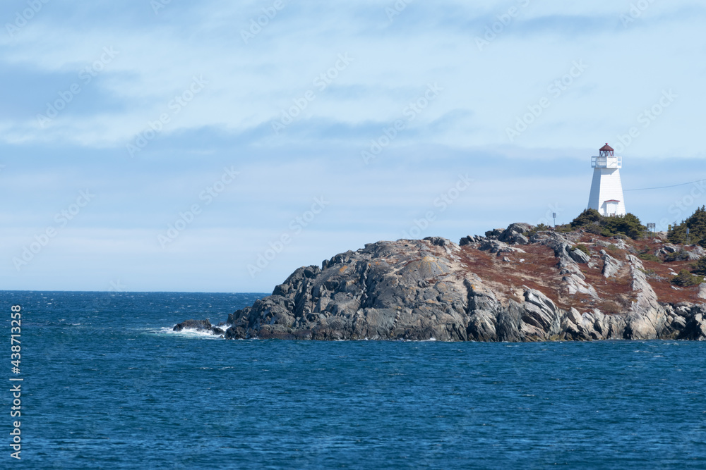 A rugged coastline and cliff in the foreground near a deep blue ocean. It's a bright sunny day with a blue sky and few clouds. There's an island in the background with greenery, buildings, and rock. 