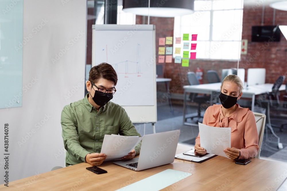 Diverse male and female colleague in face masks sitting at table with laptop reading paperwork Diverse male and female colleague in face masks sitting at table with laptop reading paperwork