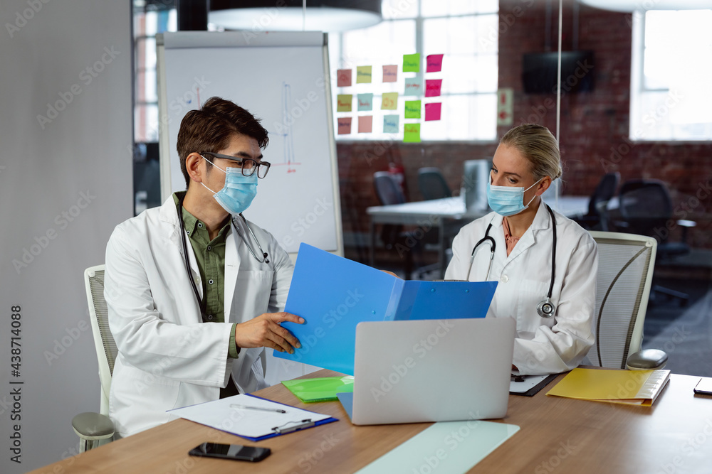 Diverse male and female doctor wearing face masks sitting in hospital office discussing paperwork Diverse male and female doctor wearing face masks sitting in hospital office discussing paperwork