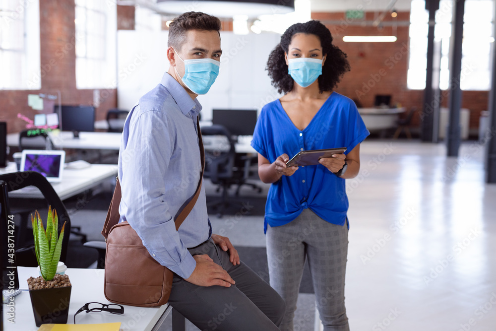 Fototapeta premium Portrait of diverse male and female colleague in face masks standing in office with tablet