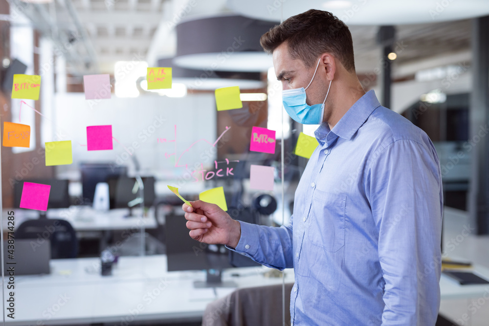 Caucasian businessman wearing face mask brainstorming, reading memo notes on wall standing in office Caucasian businessman wearing face mask brainstorming, reading memo notes on wall standing in office