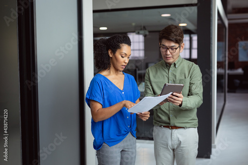 Diverse male and female colleague looking at paperwork and tablet and discussing standing in office