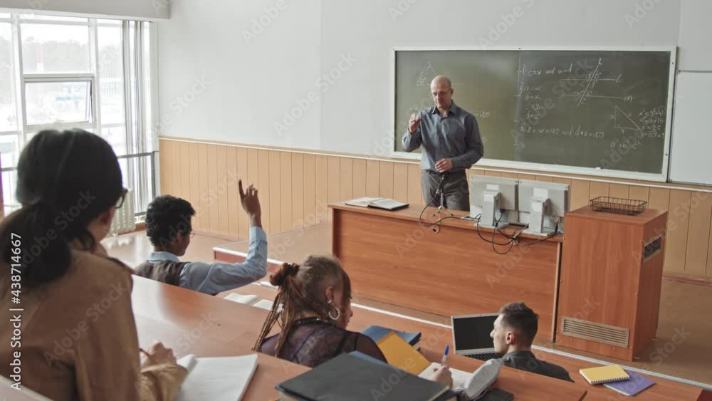 High-angle rear-view slowmo of excellent male student raising his hand to answer professor question at Maths lesson in big lecture hall High-angle rear-view slowmo of excellent male student raising his hand to answer professor question at Maths lesson in big lecture hall