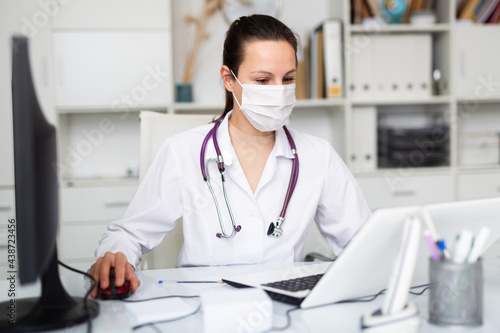 Young female doctor in a protective mask, working in the therapeutic department of the clinic, sits at a desktop in front ..of a laptop in the office