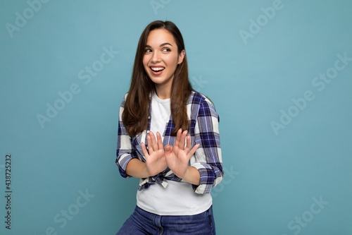 Young positive smiling beautiful brunette woman with sincere emotions wearing trendy check shirt standing isolated on blue background with empty space and showing stop gesture saying no