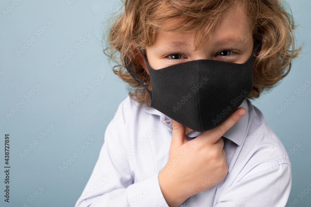 Closeup photo of handsome curly blonde little boy in medical black mask standing isolated over blue background. Coronavirus concept Closeup photo of handsome curly blonde little boy in medical black mask standing isolated over blue background. Coronavirus concept