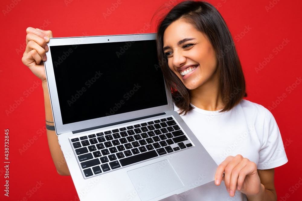 Photo of funny smiling Beautiful happy overjoyed young woman with short brunet hair holding computer laptop looking to the side wearing white t-shirt and jeans isolated over red wall background Photo of funny smiling Beautiful happy overjoyed young woman with short brunet hair holding computer laptop looking to the side wearing white t-shirt and jeans isolated over red wall background
