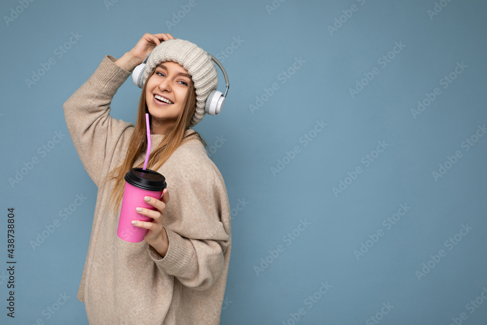 Side-profile photo of Beautiful happy smiling young blonde woman wearing beige winter sweater and hat isolated over blue background drinking beverage wearing white wireless bluetooth headsets Side-profile photo of Beautiful happy smiling young blonde woman wearing beige winter sweater and hat isolated over blue background drinking beverage wearing white wireless bluetooth headsets