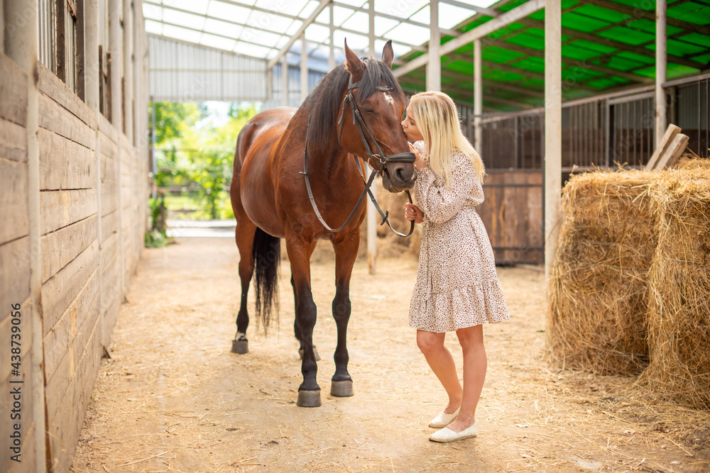 A young rider woman blonde with long hair in a dress posing with brown horse inside light stable, Russia A young rider woman blonde with long hair in a dress posing with brown horse inside light stable, Russia