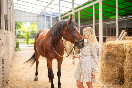 A young rider woman blonde with long hair in a dress posing with brown horse inside light stable, Russia