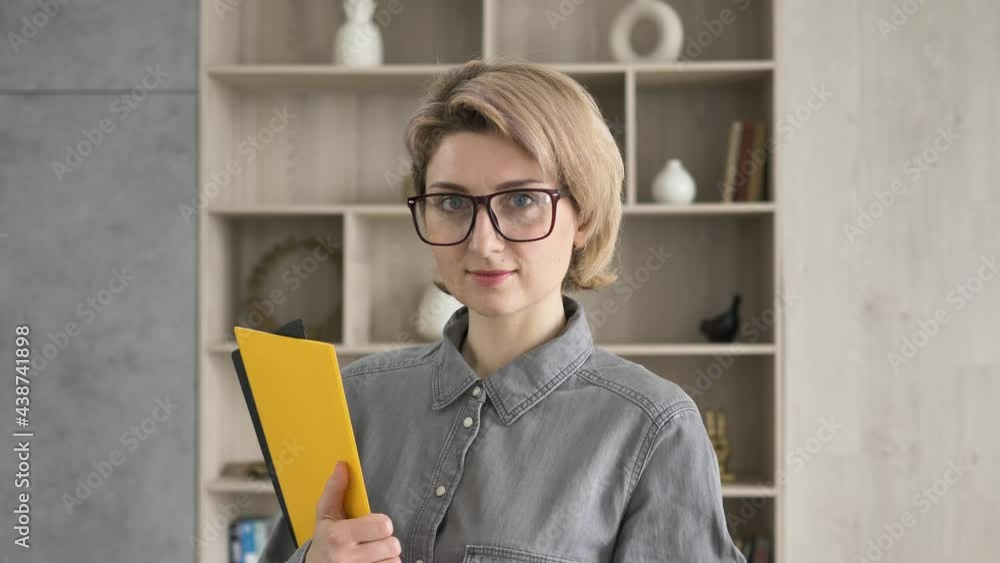 Serious lady teacher with short fair hair and glasses looks straight posing with folders against wooden shelves close view Serious lady teacher with short fair hair and glasses looks straight posing with folders against wooden shelves close view