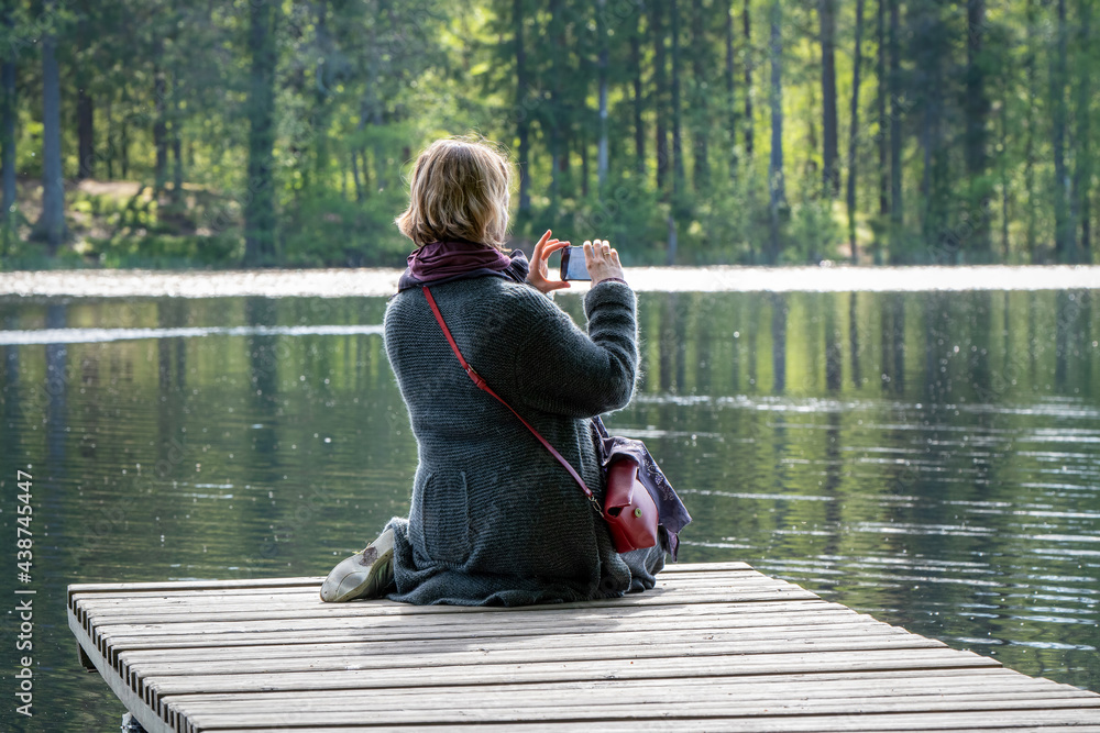 A woman takes photos with a smartphone sitting on the wooden pier on a forest lake on a sunny summer day. A woman takes photos with a smartphone sitting on the wooden pier on a forest lake on a sunny summer day.