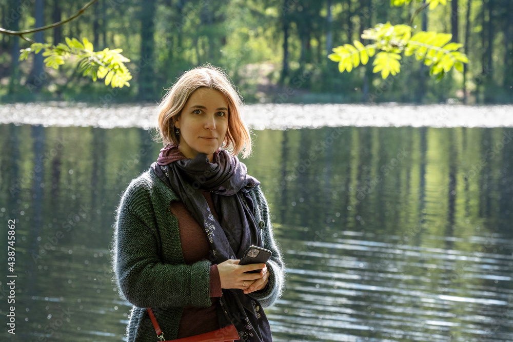 Smiling young woman standing on the shore of a forest lake on a sunny summer day.