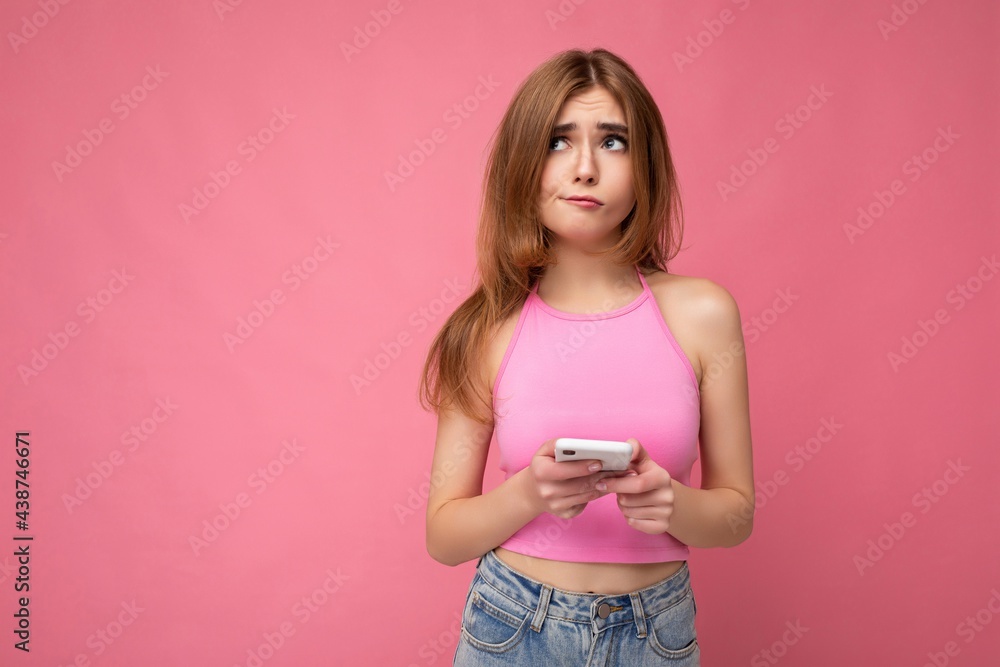 Beautiful thoughtful young blonde woman wearing pink top poising isolated on pink background with empty space holding in hand and using mobile phone surfing on the internet online looking to the side Beautiful thoughtful young blonde woman wearing pink top poising isolated on pink background with empty space holding in hand and using mobile phone surfing on the internet online looking to the side