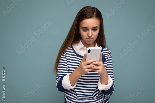 Photo shot of attractive concenrated positive good looking girl wearing casual stylish outfit poising isolated on background with empty space holding in hand and using mobile phone messaging sms