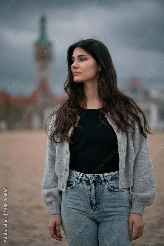 Portrait of long brown haired girl standing alone on the sand beach during cloudy weather. Spring or autumn vacation on the seaside concept. Close up portrait of caucasian woman.