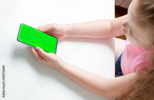 A little girl holds a smartphone with a blank green screen in two hands on a white background. Side view, copy space.