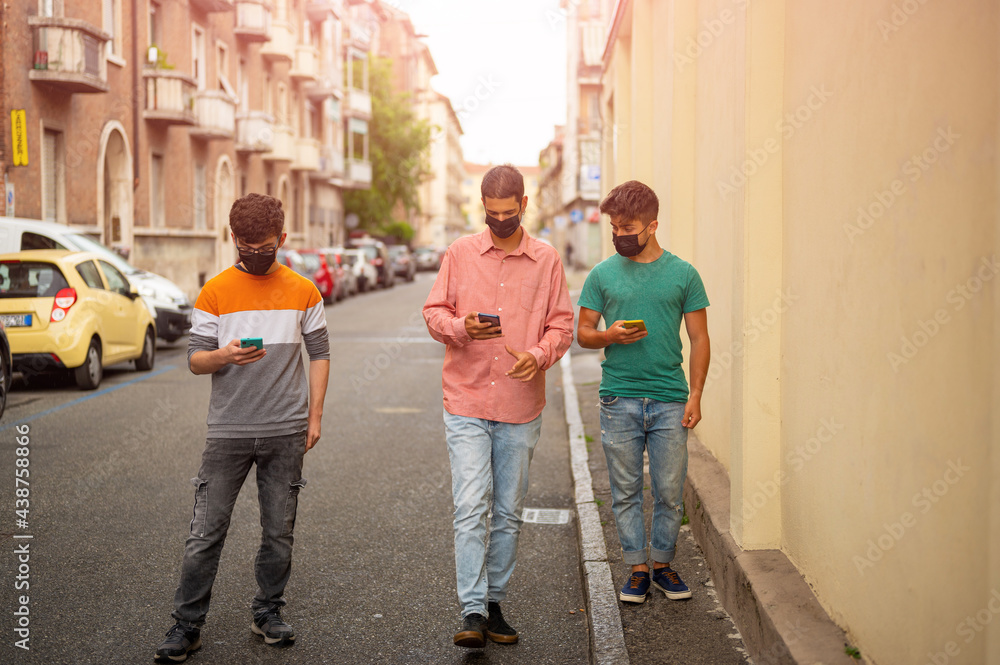 Fototapeta premium Friends group walking on city street with protective black face mask using the smartpjone. Pandemic time. New normal lifestyle concept with young people covered by protective mask. Blurred background.