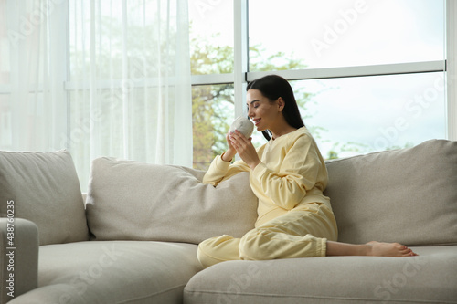 Young woman with cup of drink relaxing on sofa at home, space for text