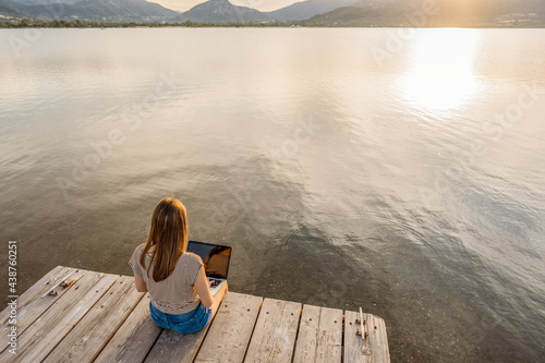 Lonely girl expressing her creativeness writing romance tales book on a wooden pier or jetty at sunset or dawn. New job opportunity at modern times using laptop and wifi internet connection technology