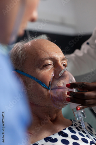 Side view of senior patient breathing assisted by respiratory tube in hospital intesive care. African doctor and nurse helping old man to breath sing face mask. photo