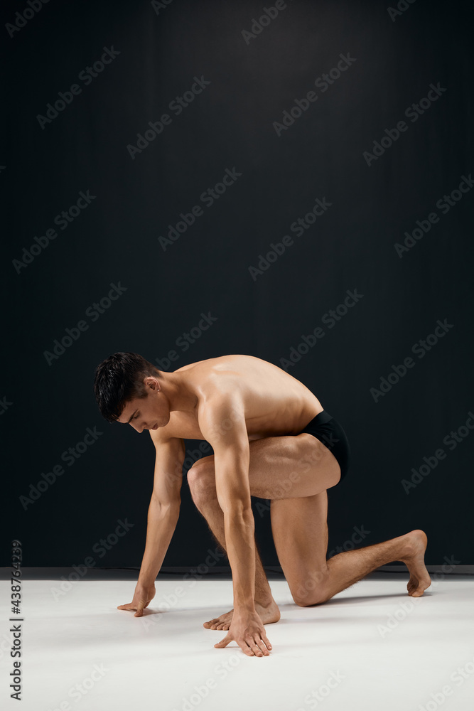 man muscularly muscled in black panties stands on his knee dark background