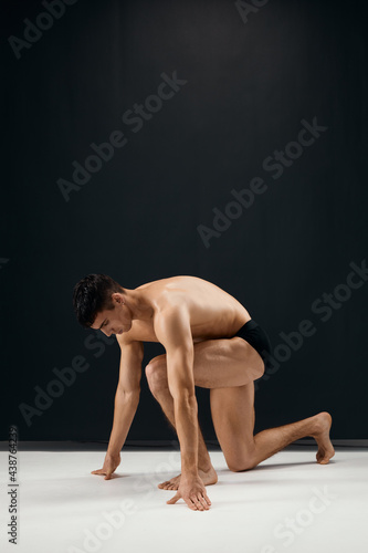 man muscularly muscled in black panties stands on his knee dark background