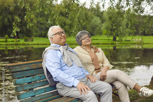 Happy married senior couple enjoying good time together. Relaxed mature man and woman sitting on an old wooden bench under a willow tree on a quiet bank of a lake or river