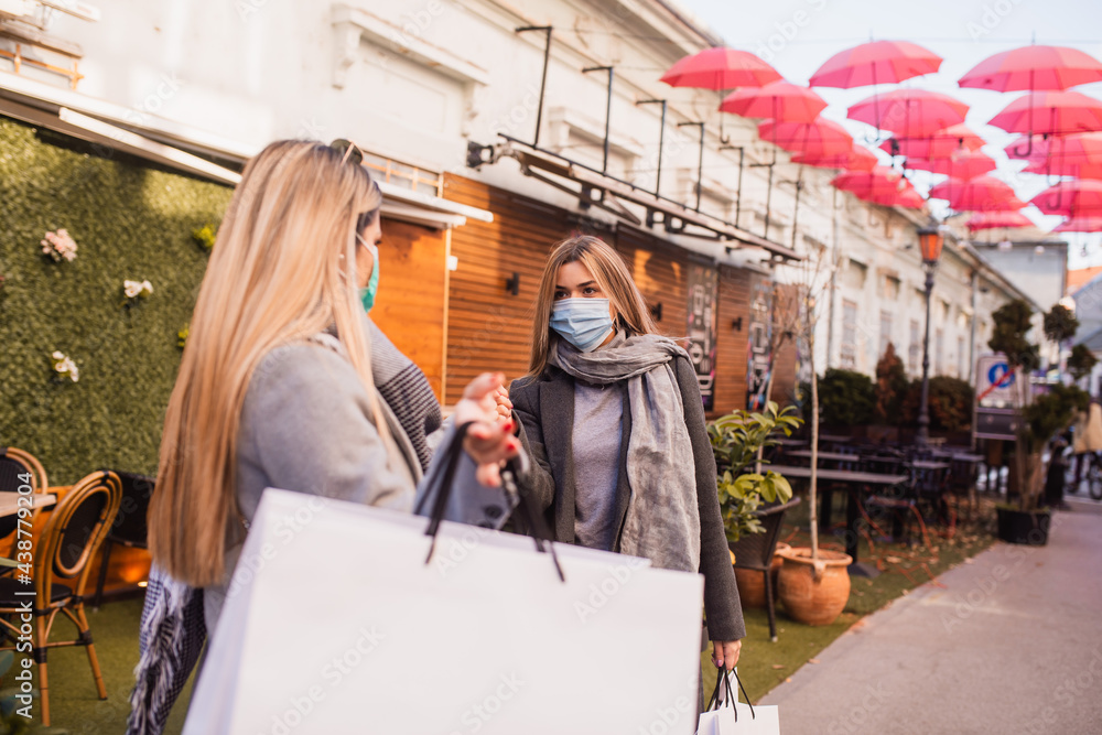 Two beautiful young Caucasian women with a face mask greet each other on the street. Shopping during the COVID-19 coronavirus pandemic Two beautiful young Caucasian women with a face mask greet each other on the street. Shopping during the COVID-19 coronavirus pandemic