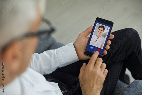 Dating online. Senior man likes the photo of a young beautiful laughing european woman. A man in a white shirt sits on a sofa and looks at a photo on a digital smartphone, puts a like on the photo