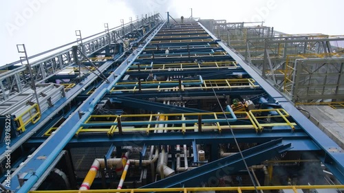 Camera movement from the base of the refinery tower. The complex structure of the metal construction and the cloudy sky with clouds of steam are visible.